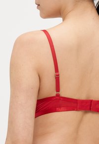 Raudonos spalvos apatinių kelnaičių liemenėlė su reguliuojamomis petnešėlėmis, atlasine tekstūra ir užsegimu gale su kabliukais. Lygus dizainas su minimaliais akcentais.