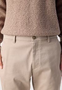 Beige Chinos mit glatter Textur, ausgestattet mit einem Knopfverschluss und seitlichen Taschen, kombiniert mit einem strukturierten, hellbraunen Pullover.