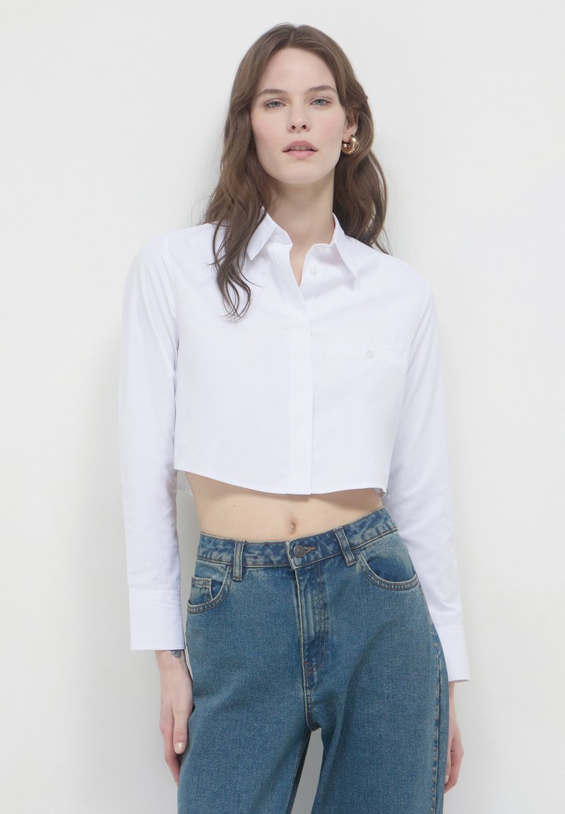 Donna con lunghi capelli castani che indossa orecchini a cerchio dorati, una camicia bianca corta con bottoni e jeans blu a vita alta, contro uno sfondo bianco semplice.