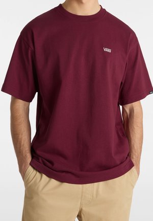 Mann trägt ein schlichtes weinrotes Vans-T-Shirt mit kurzen Ärmeln und beigen Hosen, Hände in den Taschen, vor schlichtem Hintergrund.