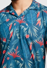 Camisa de manga corta en tela azul, con un diseño tropical que presenta grandes hojas verdes, flores rojas y acentos. Con cuello y un corte relajado.