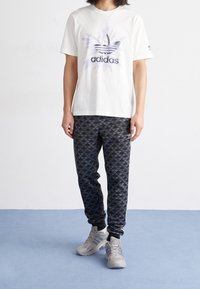 Bílý bavlněný tričko s purpurovým logem Adidas, doplněné černými vzorovanými tepláky a šedými teniskami s měkkou podrážkou.