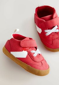 Zapatillas de tela rojas con acentos y cordones blancos, correa de velcro y suelas de goma marrón que presentan un patrón de agarre texturizado.