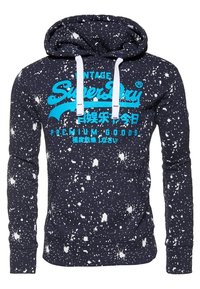 Granatowa bluza z kapturem z nadrukiem w białe plamy farby, wyrazisty turkusowy logo i tekst "SuperDry" oraz białe sznurki do ściągania.