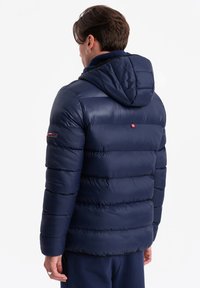 Mörkblå puffjacka med huva, som har kviltade sektioner, slät textur och en logodetalj på ärmen.