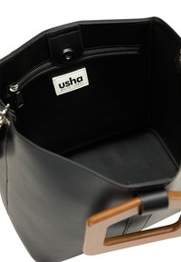 Bolso de mano de cuero negro con una textura suave, que presenta un asa cuadrada de madera y un bolsillo con cremallera en el interior. Etiqueta interior visible.
