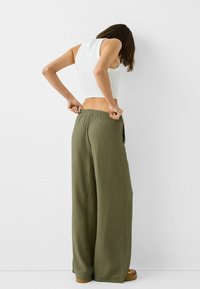 Volné olivově zelené kalhoty vyrobené z lehkého materiálu, s elastickým pasem a širokým střihem nohavic. Model má na sobě bílý top bez rukávů.