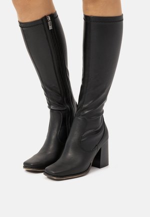 Bottes noires en cuir jusqu'au genou avec un bout carré, fermeture éclair latérale et talon épais. Le design présente des coutures pour ajouter de la texture.