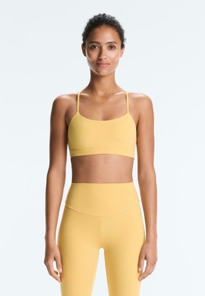 Kobieta stojąca, ubrana w żółty sportowy biustonosz i legginsy z wysokim stanem, o neutralnym wyrazie twarzy i kręconych włosach zaczesanych do tyłu.