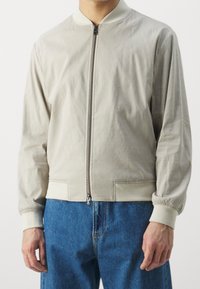 Světle šedá bomber bunda se zipem vpředu, vroubkovaným límcem a manžetami a pružným lemem. Hladká tkanina s minimálními švy.