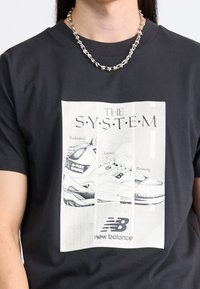 Černé tričko s bílým obdélníkovým grafikou tří tenisek New Balance a textem "SYSTÉM" a logem "new balance".
