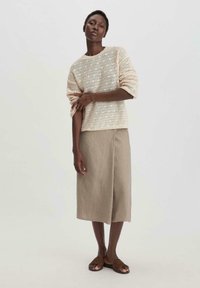 Helles beigefarbenes Strickoberteil mit offenem Webmuster, kombiniert mit einem hellbraunen Leinen-Midirock. Das Model trägt braune flache Sandalen. Einfaches und zurückhaltendes Design.