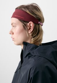 Maroon athletic headband with "Peak Performance" logo. Smooth fabric, elastic at the back for adjustment. Partially visible black jacket.