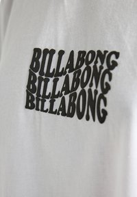 T-shirt bianca con un logo "BILLABONG" ripetuto in nero di dimensioni variabili, caratterizzato da un font audace e arrotondato sulla parte anteriore.