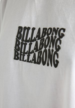 T-shirt bianca con un logo "BILLABONG" ripetuto in nero di dimensioni variabili, caratterizzato da un font audace e arrotondato sulla parte anteriore.