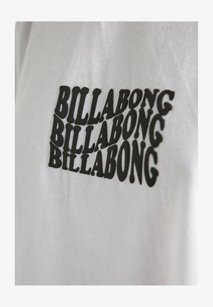 Witte T-shirt met een herhaald zwart "BILLABONG" logo in verschillende groottes, met een vetgedrukt, rond lettertype aan de voorkant.