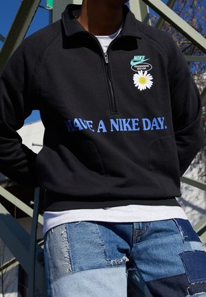 Person trägt einen schwarzen Nike Pullover mit dem Text "Have a Nike Day" und Gänseblümchen-Grafik, kombiniert mit Patchwork-Blue-Denim-Jeans im Freien.