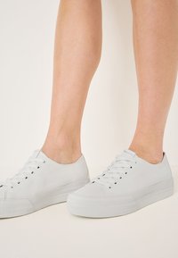 Weiße niedrige Sneakers aus Canvas mit runder Zehenpartie, flacher Schnürung und strukturiertem Gummi-Sohle. Keine sichtbaren Muster oder Akzente.