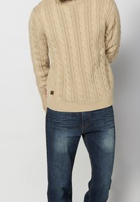Pull beige tricoté avec motif torsadé, manches longues et ourlet côtelé, associé à un jean en denim bleu foncé avec un léger effet délavé.