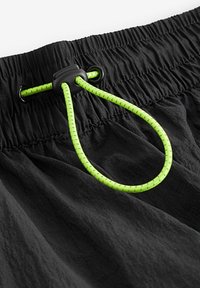 Mustad mustri elastse vööga lühikesed püksid, millel on erks roheline pael, mis on valmistatud tekstuursest materjalist ja kinnitatud musta plastikust klambriga.