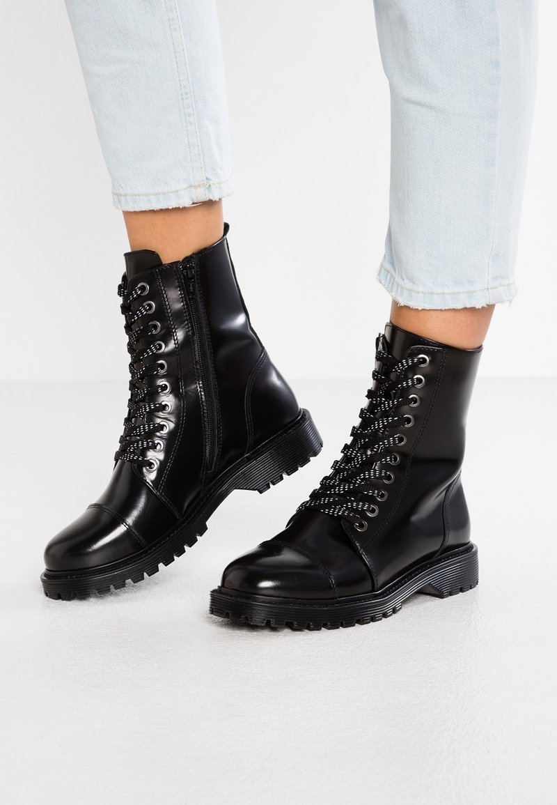 Botas de tobillo negras brillantes con cordones y cremallera lateral, combinadas con jeans cortos azul claro sobre una superficie blanca.