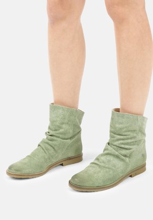 Onderbenen die groene suède enkellaarsjes dragen met platte bruine zolen op een witte achtergrond.