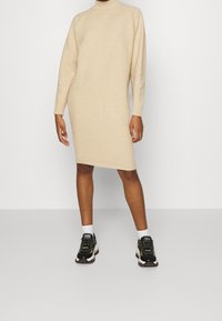 Abito lavorato a maglia beige con colletto alto e maniche lunghe, caratterizzato da una silhouette dritta e polsini a costine, abbinato a sneakers atletiche nere.