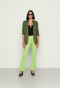 Camisola preta, blazer verde em padrão escocês, calças verde em corte flare e sandálias de salto pretas. Óculos de sol elegantes completam o visual.