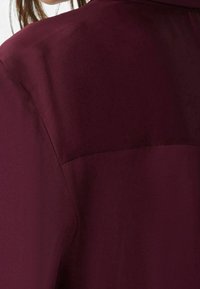 Vinrød bluse med et glat, glinsende stof, skjult knapliste og en skulderpasse med detaljer bagpå for et diskret design.