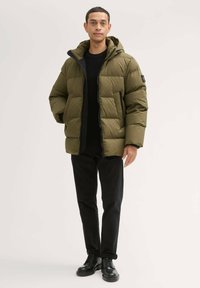 Oliwkowa puchowa kurtka z zapięciem na zamek, kapturem i czarnymi akcentami; zestawiona z czarnymi spodniami i czarnymi butami. Posiada pikowaną teksturę.