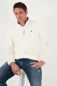 Valge fliispusa poolse tõmblukuga, millel on rindkeres väike must logo, koos siniste kulunud teksadega.