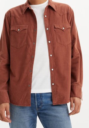 Mand iført en rustfarvet kordfløjls skjorte over en hvid t-shirt og blå jeans, stående mod en ensfarvet baggrund.