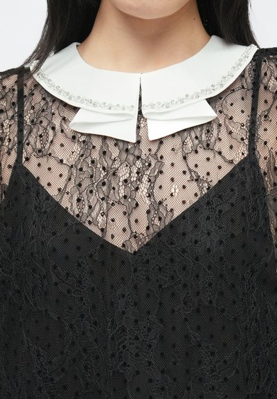 Svart blonderkjole med et gjennomsiktig polkadottlag, med hvit krage pyntet med dekorative elementer og V-formet utringning.