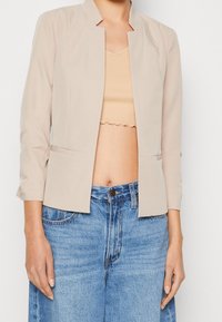 Blazer beige con cuello mandarín, diseño estructurado y textura suave. Combinado con un top beige corto y jeans de mezclilla azul de talle alto.