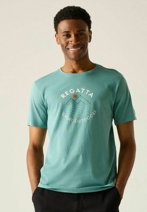 Hombre sonriente con camiseta color teal con gráfico de montaña y olas y el texto "Regatta Great Outdoors", con las manos en los bolsillos.