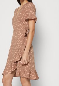 Vestido envolvente marrón con lunares, con mangas cortas con volantes, que presenta un dobladillo con volantes y un detalle de lazo en la cintura, hecho de una tela ligera.