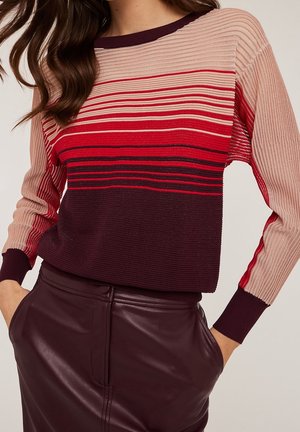 Femme portant un pull en maille côtelée à rayures horizontales beige, rouge et bordeaux, assorti à un pantalon en cuir bordeaux foncé.