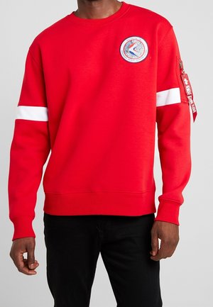 Sweat-shirt rouge avec un patch logo rond, des rayures blanches sur les manches et une poche zippée avec une étiquette en tissu sur le bras gauche.