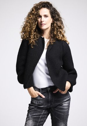 Frau mit lockigem Haar, die eine strukturierte schwarze Jacke, ein weißes Hemd und verblasste schwarze Jeans trägt. Sie steht mit den Händen in den Taschen vor einem hellen Hintergrund.