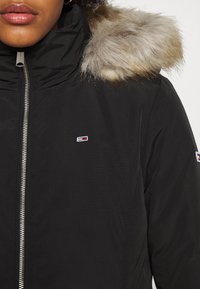 Veste noire avec une fermeture éclair à l'avant et un bord en fausse fourrure sur la capuche. Dispose d'un petit logo sur la poitrine et d'un tissu texturé.