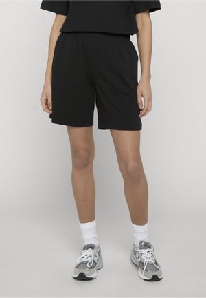Person iført sorte shorts og sort top, hvide sokker og grå løbesko, står foran en ensfarvet lys baggrund.