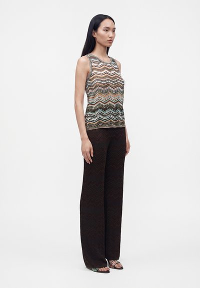 Femme debout portant un top sans manches à motifs et un pantalon ample foncé avec des sandales ouvertes, sur un fond blanc uni.