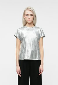 Kvinde med glat blond hår iført en skinnende sølvkortærmet top og sorte bukser, der står foran en ensfarvet lys baggrund.