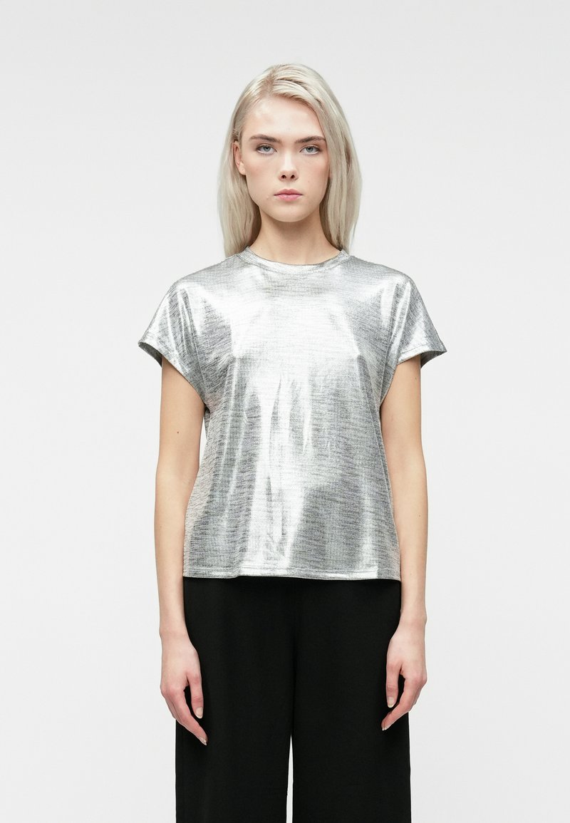 Kvinde med glat blond hår iført en skinnende sølvkortærmet top og sorte bukser, der står foran en ensfarvet lys baggrund.