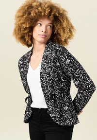 Chaqueta con estampado floral en blanco y negro, con cintura ceñida, tejido ligero, apertura frontal y dos lazos en la cintura. Llevada sobre una blusa blanca.