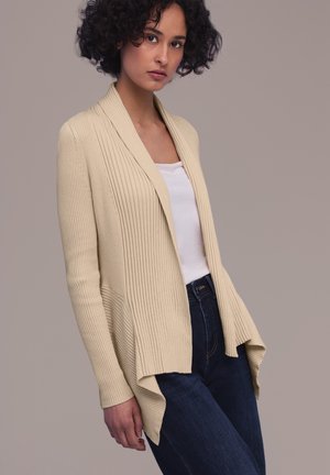 Femme portant un cardigan crème à côtes sur un haut blanc et un jean bleu foncé, debout devant un fond beige uni.