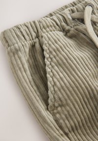 Pantalon de survêtement en velours côtelé de couleur olive claire avec texture côtelée, taille élastique et cordon de serrage ajustable. Dispose de poches latérales.