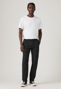 Mand med fletning bærer en simpel hvid t-shirt, sorte bukser og sort-hvide sneakers, stående med hænderne i lommerne.