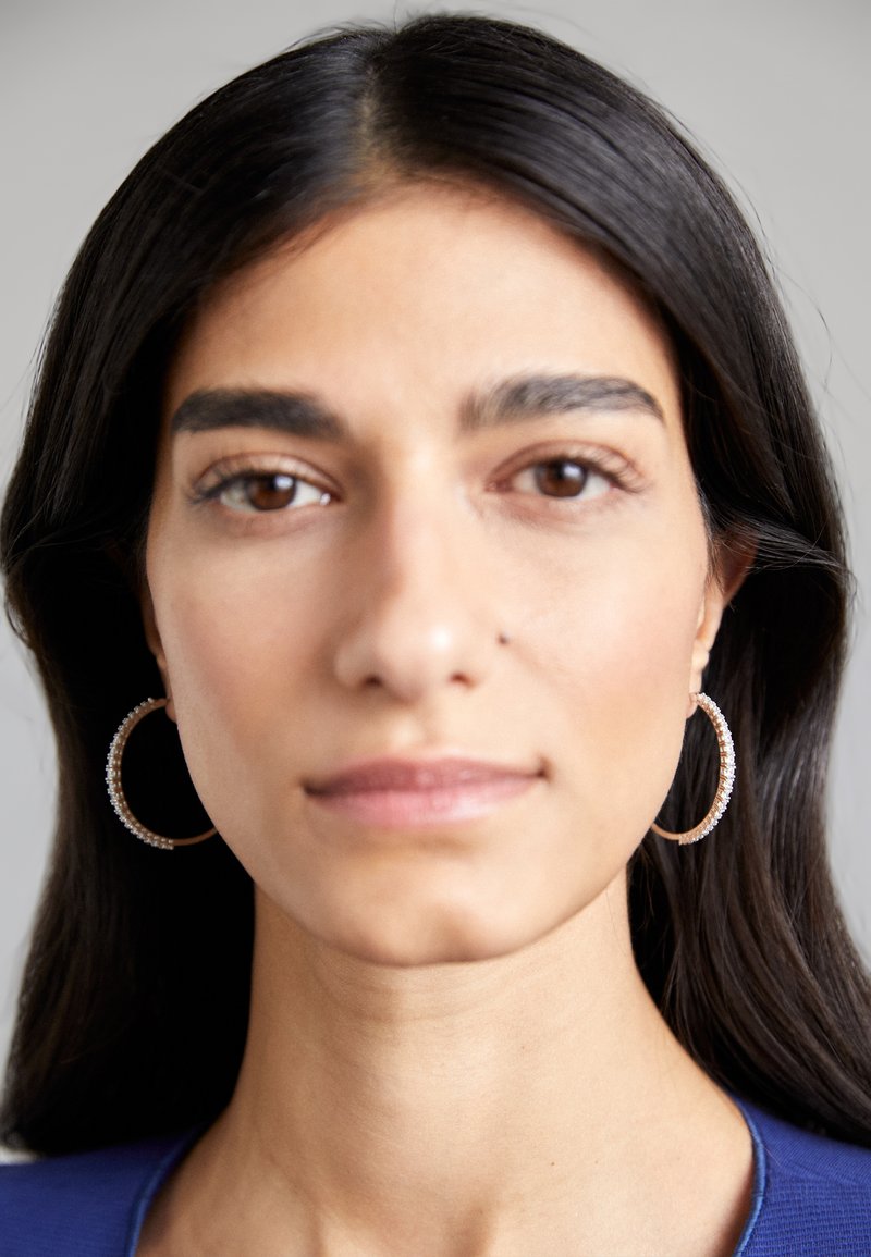 Boucles d'oreilles en or en forme de créole ornées de petites pierres précieuses claires, portées par une personne aux cheveux longs et foncés et portant un haut bleu, sur un fond neutre.