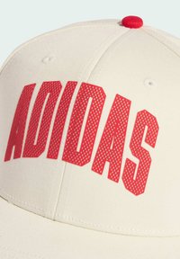 Gorra de béisbol crema con una forma estructurada, que presenta el texto en rojo "ADIDAS" en un diseño perforado y un botón superior rojo.